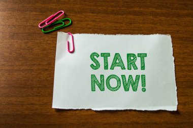 Word writing text Start Now. Business concept for do not hesitate get working or doing stuff right away Close up blank stationary paper hold three colored clips lying wooden table.