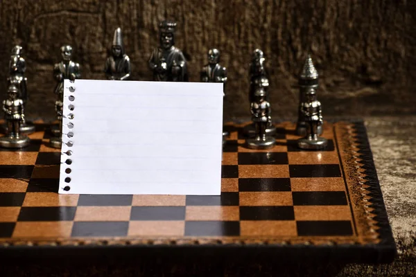 На фоне шахматной доски перед шахматами черной тематики оторван и сложен чистый лист с чистым спиральным шнуром — стоковое фото