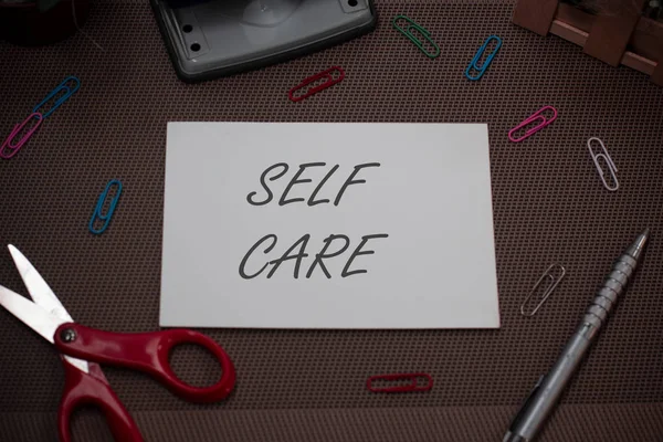 Tulisan tangan menulis Self Care. Konsep yang berarti praktik mengambil tindakan untuk meningkatkan satu s adalah gunting kesehatan sendiri dan alat tulis ditambah lembaran polos di atas latar belakang bertekstur . — Stok Foto