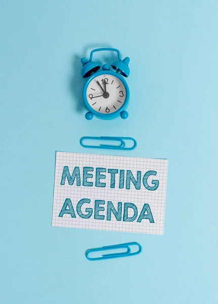 Handwriting text writing Meeting Agenda. Concept meaning items that participants hope to accomplish at a meeting Alarm clock wakeup squared blank paper sheet clips colored background.