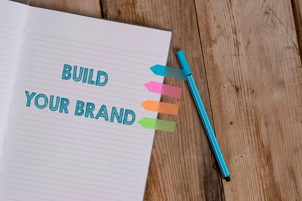 Conceptual hand writing showing Build Your Brand. Business photo text enhancing brand equity using advertising campaigns Striped notebook colored arrow banners ballpoint wooden background. — Stock Photo, Image