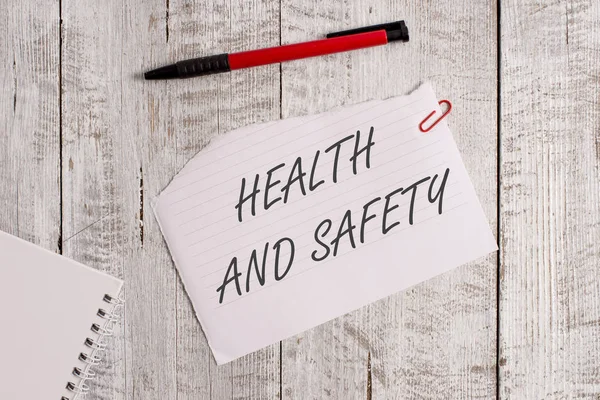 Woord tekst schrijven gezondheid en veiligheid. Bedrijfsconcept voor procedures die bedoeld zijn om ongevallen te voorkomen op werkplek gescheurd papier en notebookvel met een pen bovenop een houten tafel. — Stockfoto