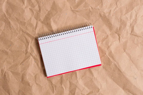 Skrivbord av skrynkliga hantverk papper med en öppen fyrkantig papper spiral röd anteckningsbok och kontorsmaskiner. Pappersbord med papper och studietillbehör. — Stockfoto