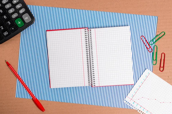 Mesa de papelão com uma folha azul listrada, papel notebook e eletrodomésticos de escritório. Tabela de papelão com diferentes tamanhos e tipos de papéis, um gráfico com números e materiais de estudo. — Fotografia de Stock