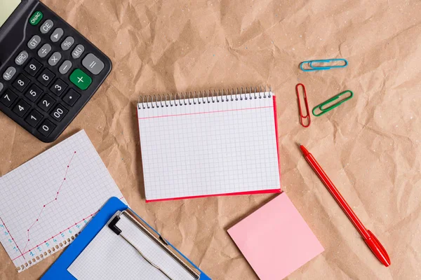 Desk made of crumpled craft paper with an open square paper spiral red notebook and office appliances. Papercraft table with papers and study supplies.