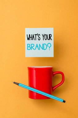 Marka Sorunuz Nedir'i gösteren not yazma. İş fotoğraf showcasing ürün logosu hakkında soran yapar ya da ne iletişim Ön görünüm kahve fincanı renkli yapışkan not kalem sarısı renkli arka plan.