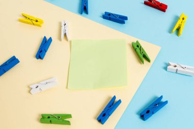Mavi arka plan ve sarı kare ortasında boş dikdörtgen şeklinde hatırlatma notu etrafında yerleştirilen renkli clothespin. Ofis malzemeleri, bir bağlantı elemanı tarafından sabitlenmiş boş kağıt.