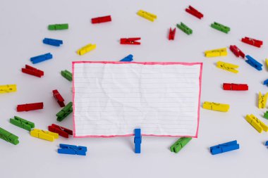 Beyaz bir zeminin ortasında boş dikdörtgen kare şeklinde hatırlatma notu etrafında yerleştirilen renkli clothespin. Ofis malzemeleri, bir bağlantı elemanı tarafından sabitlenmiş boş kağıt.