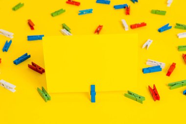 Sarı bir arka plan ortasında boş dikdörtgen kare şeklinde hatırlatma notu etrafında yerleştirilen renkli clothespin. Ofis malzemeleri, bir bağlantı elemanı tarafından sabitlenmiş boş kağıt.
