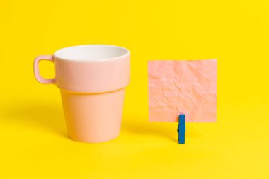 Somon bardağı ve mavi bir çamaşır iğnesi içinde boş bir not kağıdı. Kare dikdörtgen şeklinde renkli anımsatıcı sarı bir arka plan. Boş alan ve ofis malzemeleri.