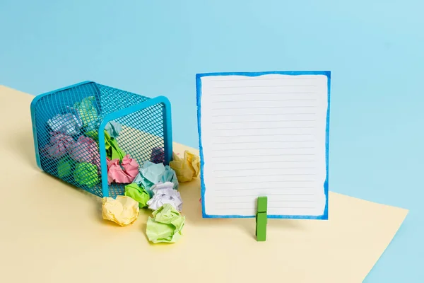 Pequeno caixote do lixo cheio de papel colorido amassado e um pano pendurado uma nota de papel colorido em um fundo amarelo e azul. Material de escritório e lembrete vazio . — Fotografia de Stock