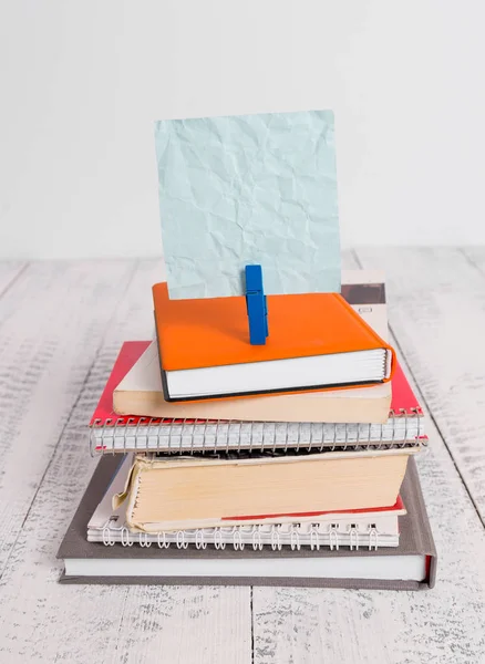 Square shaped paper above a pile of notebooks fixed by a blue clothespin. Different books stacked under an empty reminder colorful note. White wooden floor and white background.