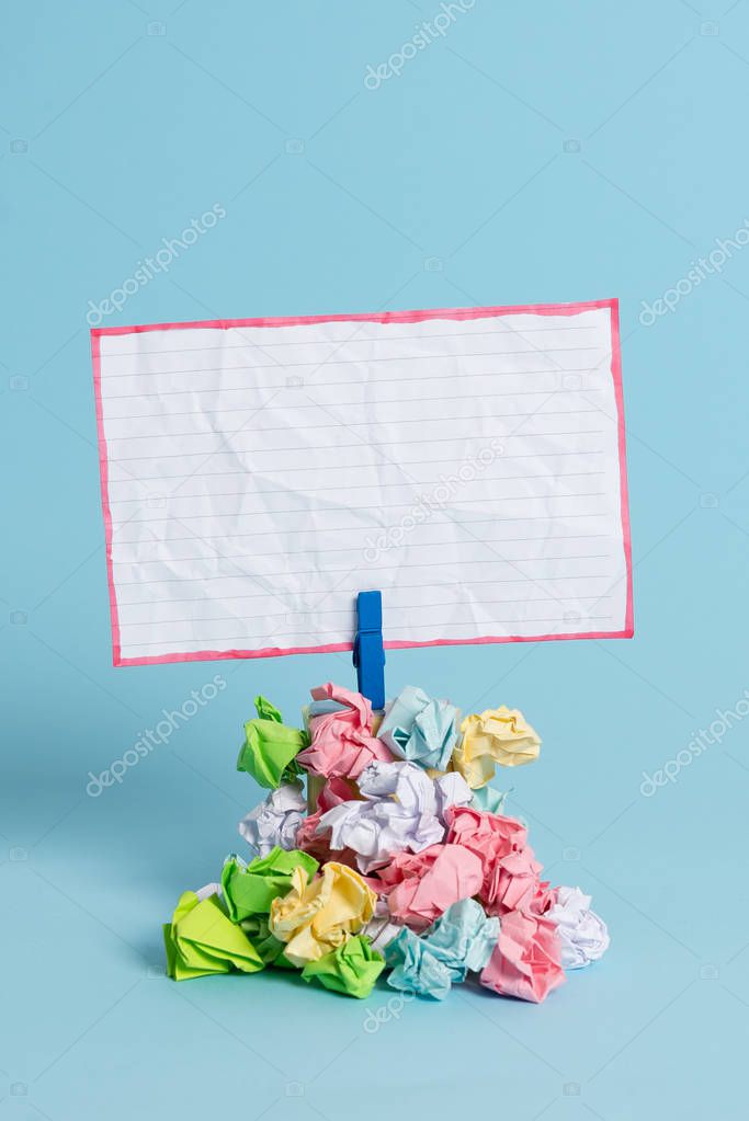 Square shaped reminder above a pile of crumpled papers fixed by a blue clothespin. Different sheets stacked under an empty colorful note. Light blue background.