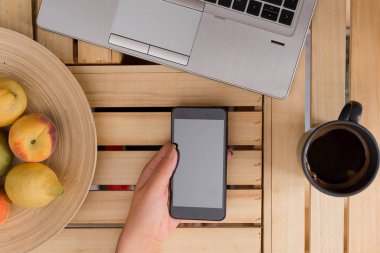 Genç bayan kullanarak ve siyah kahve bir fincan ile bir sandık masada karanlık bir akıllı telefon tutarak. Ofis malzemeleri, cep telefonu, teknolojik cihazlar ve ahşap masa.