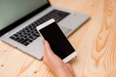 Genç bayan sol eliyle akıllı telefon tutuyor. Siyah klavye, kahve, ofis malzemeleri, teknolojik cihazlar ve ahşap masa ile arka planda Gri dizüstü bilgisayar.