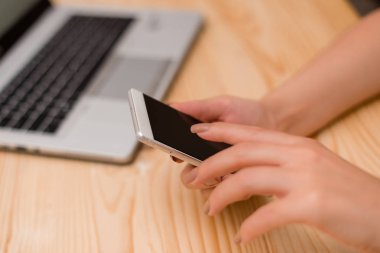 Genç bayan akıllı telefon kullanıyor ve ekrana dokunuyor. Siyah klavye ve bir fincan kahve ile arka planda gri dizüstü bilgisayar. Ofis malzemeleri, teknolojik cihazlar ve ahşap masa.