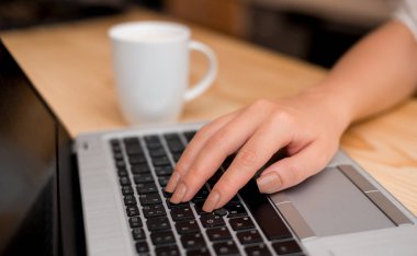 Genç bayan gri bir dizüstü bilgisayar kullanarak ve arka planda kahve veya çay bir fincan ile siyah klavye yazarak. Ofis malzemeleri, teknolojik cihazlar ve ahşap masa.