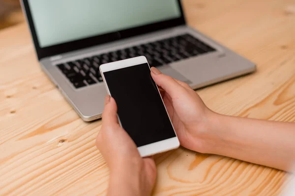 Jovencita sosteniendo un smartphone con ambas manos. Ordenador portátil gris en el fondo con teclado negro. Materiales de oficina, dispositivos tecnológicos y escritorio de madera . — Foto de Stock