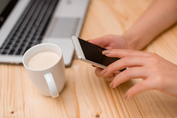 Jovencita usando un smartphone y tocando la pantalla. Ordenador portátil gris en el fondo con teclado negro y una taza de café. Materiales de oficina, dispositivos tecnológicos y escritorio de madera . — Foto de Stock