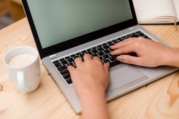 Молода леді використовує сірий ноутбук і друкує в чорній клавіатурі з чашкою кави або чаю на задньому плані. Офісне приладдя, технологічні пристрої та дерев'яний стіл . — стокове фото