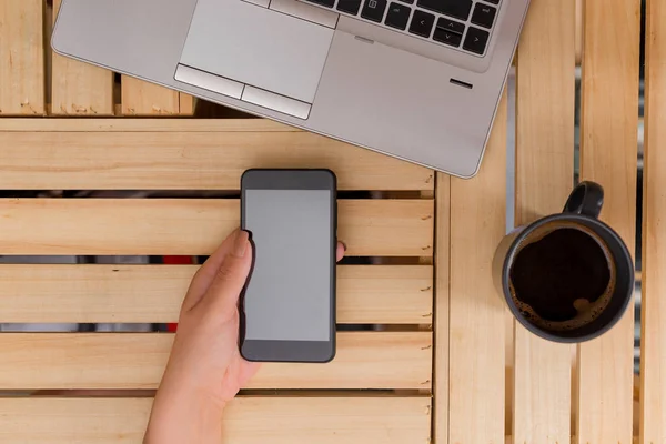 Νεαρή κυρία χρησιμοποιώντας και κρατώντας ένα σκοτεινό smartphone σε ένα κιβώτιο τραπέζι με μια κούπα του μαύρου καφέ. Αναλώσιμα γραφείου, κινητό τηλέφωνο, τεχνολογικές συσκευές και ξύλινο γραφείο. — Φωτογραφία Αρχείου