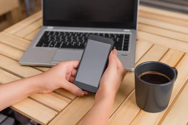 Ung dam använder och håller en mörk smartphone i en låda bord med en mugg svart kaffe. Kontorsmateriel, mobiltelefon, tekniska enheter och trä skrivbord. — Stockfoto