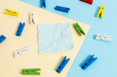 Mavi arka plan ve sarı kare ortasında boş dikdörtgen şeklinde hatırlatma notu etrafında yerleştirilen renkli clothespin. Ofis malzemeleri, bir bağlantı elemanı tarafından sabitlenmiş boş kağıt.