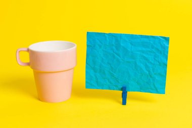 Somon bardağı ve mavi bir çamaşır iğnesi içinde boş bir not kağıdı. Kare dikdörtgen şeklinde renkli anımsatıcı sarı bir arka plan. Boş alan ve ofis malzemeleri.