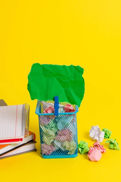Pequeno caixote do lixo cheio de papel colorido amassado e um pano azul pendurado uma nota colorida em um fundo amarelo. Material de escritório e lembrete vazio fixados em uma lata . — Fotografia de Stock