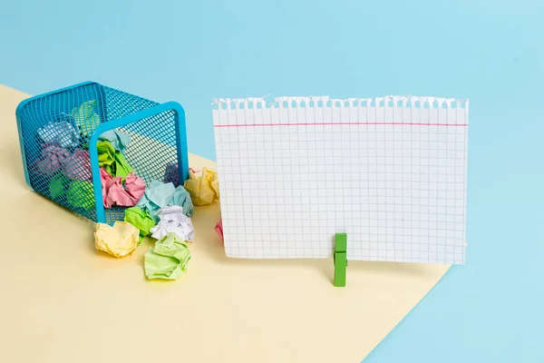 Pequeño cubo de basura con punta lleno de papel de colores arrugados y un tendedero colgando una nota de papel de colores en un fondo amarillo y azul. Materiales de oficina y recordatorio vacío . —  Fotos de Stock