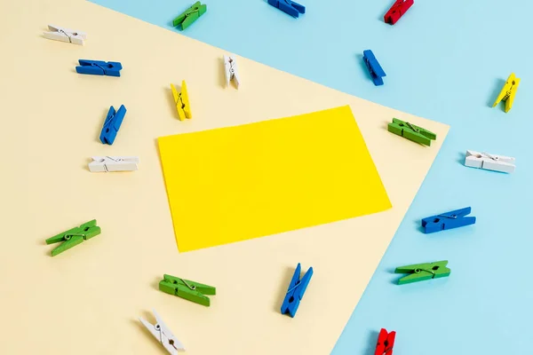 Mavi arka plan ve sarı kare ortasında boş dikdörtgen şeklinde hatırlatma notu etrafında yerleştirilen renkli clothespin. Ofis malzemeleri, bir bağlantı elemanı tarafından sabitlenmiş boş kağıt. — Stok fotoğraf