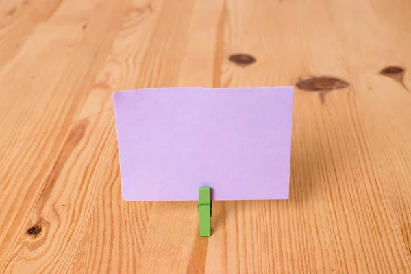 Tom rektangel fyrkantig formad påminnelse anteckning i mitten av ett trägolv som innehas av en grön klädnypa. Färgat papper i trämark med spår och springor. Kontorsmateriel. — Stockfoto