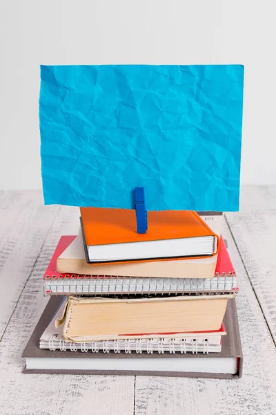 Square shaped paper above a pile of notebooks fixed by a blue clothespin. Different books stacked under an empty reminder colorful note. White wooden floor and white background.