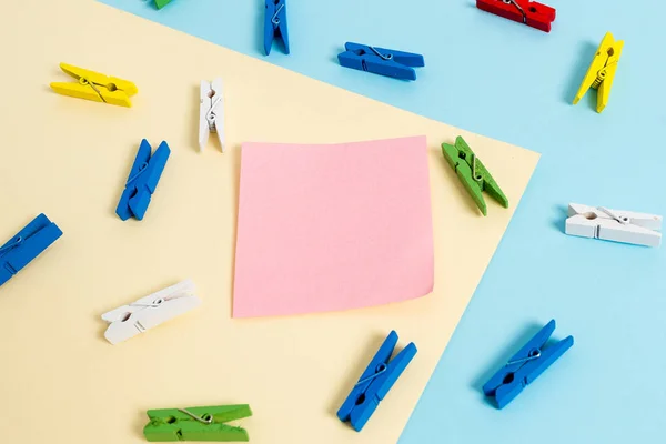 Espina de ropa de color colocada alrededor de la nota recordatorio en forma de rectángulo vacío en el centro de un fondo azul y un cuadrado amarillo. Material de oficina, papel en blanco fijado por un cierre . — Foto de Stock