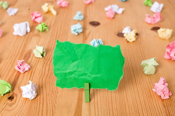 Gekleurde verfrommeld papier rond een lege rechthoek vierkant vormige herinnering notitie in het midden van een houten vloer gehouden door een groene wasknijper. Hout met groeven. Kantoorbenodigdheden. — Stockfoto