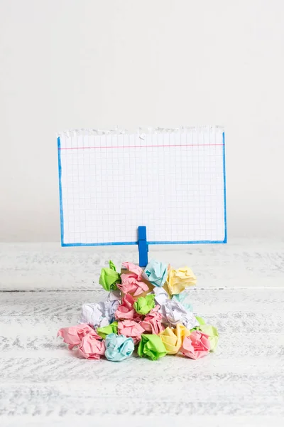 Recordatorio en forma cuadrada sobre una pila de papeles arrugados fijados por un pasador de ropa azul. Diferentes hojas apiladas bajo una nota colorida vacía. Suelo de madera blanco y fondo blanco . — Foto de Stock