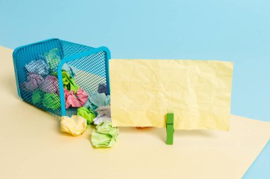 Küçük uçlu çöp kutusu buruşuk renkli kağıt ve sarı ve mavi arka planda renkli bir kağıt notasma bir clothespin dolu. Ofis malzemeleri ve boş anımsatıcı.