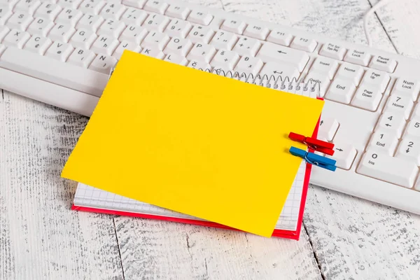 Vierkante papieren notitieboekje met een rechthoek vormige lege notitie boven vastgemaakt door twee wasknijpers op het blad. Wit toetsenbord onder een beurt en lichte houten vloer achtergrond. — Stockfoto