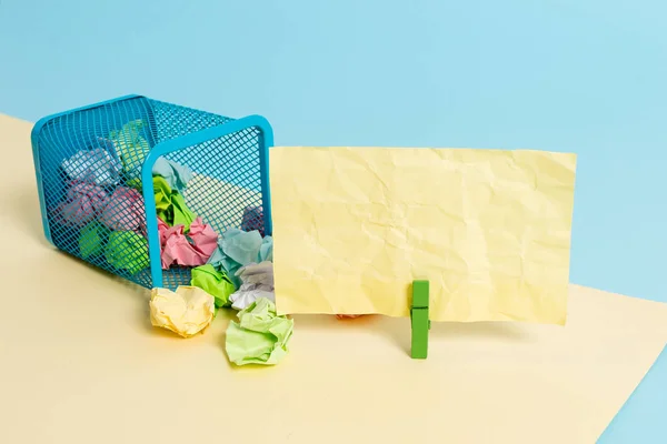 Pequeño cubo de basura con punta lleno de papel de colores arrugados y un tendedero colgando una nota de papel de colores en un fondo amarillo y azul. Materiales de oficina y recordatorio vacío . —  Fotos de Stock