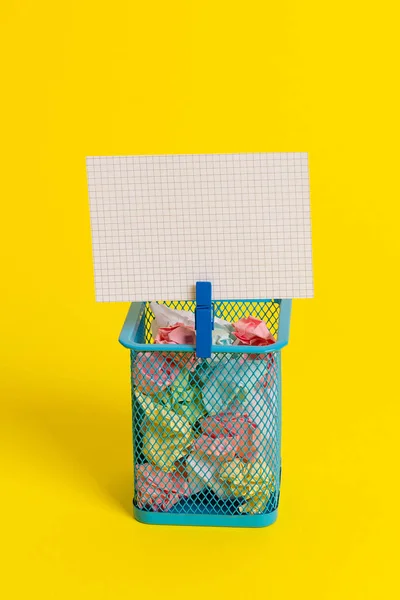 Petite poubelle pleine de papier coloré froissé et une pince à linge bleue accrochant une note colorée dans un fond jaune. Fournitures de bureau et rappel vide fixés sur une boîte . — Photo