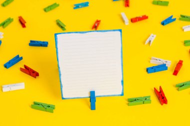 Sarı bir arka plan ortasında boş dikdörtgen kare şeklinde hatırlatma notu etrafında yerleştirilen renkli clothespin. Ofis malzemeleri, bir bağlantı elemanı tarafından sabitlenmiş boş kağıt.