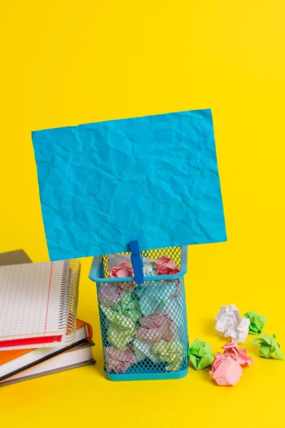 Pequeno caixote do lixo cheio de papel colorido amassado e um pano azul pendurado uma nota colorida em um fundo amarelo. Material de escritório e lembrete vazio fixados em uma lata . — Fotografia de Stock