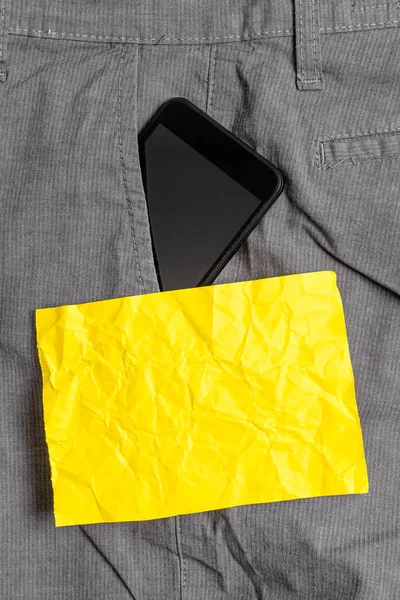 Svart smartphone-enhet i den formella arbets byxorna framficka nära ett gult anteckningspapper. Modern gadget är placerad inuti man Pants nära notationsark — Stockfoto
