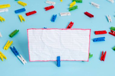 Mavi bir arka plan ortasında boş dikdörtgen kare şeklinde hatırlatma notu etrafında yerleştirilen renkli clothespin. Ofis malzemeleri, bir bağlantı elemanı tarafından sabitlenmiş boş kağıt.