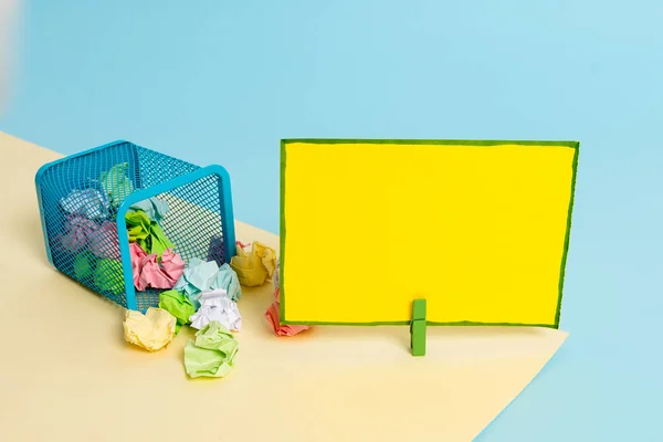 Little tipped trash bin full of crumpled colored paper and a clothespin hanging a colorful paper note in a yellow and blue background. Office supplies and empty reminder.