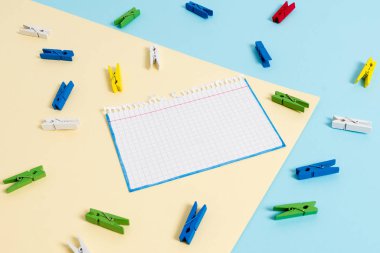 Mavi arka plan ve sarı kare ortasında boş dikdörtgen şeklinde hatırlatma notu etrafında yerleştirilen renkli clothespin. Ofis malzemeleri, bir bağlantı elemanı tarafından sabitlenmiş boş kağıt.