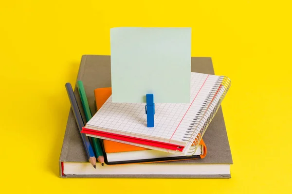Books, pencils and an empty rectangle square shaped reminder above a blank opened notebook held by a blue clothespin. Office supplies in a yellow background. — Stock Photo, Image