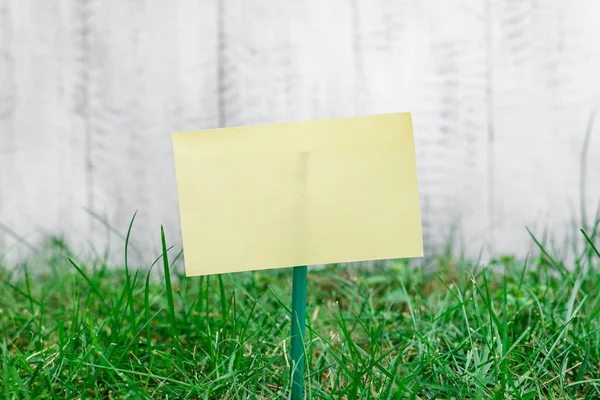 Plain paper cardboard attached to a stick and plugged in the grassy land. Yellow empty sheet is placed in ground with green grass. Photography idea with small object