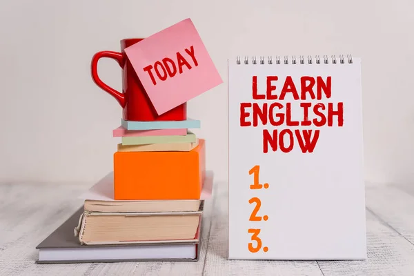 Word writing text Learn English Now. Business concept for gain or acquire knowledge and skill of english language Cup sticky note stacked note pads books square box spiral wooden table.