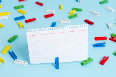 Mavi bir arka plan ortasında boş dikdörtgen kare şeklinde hatırlatma notu etrafında yerleştirilen renkli clothespin. Ofis malzemeleri, bir bağlantı elemanı tarafından sabitlenmiş boş kağıt.